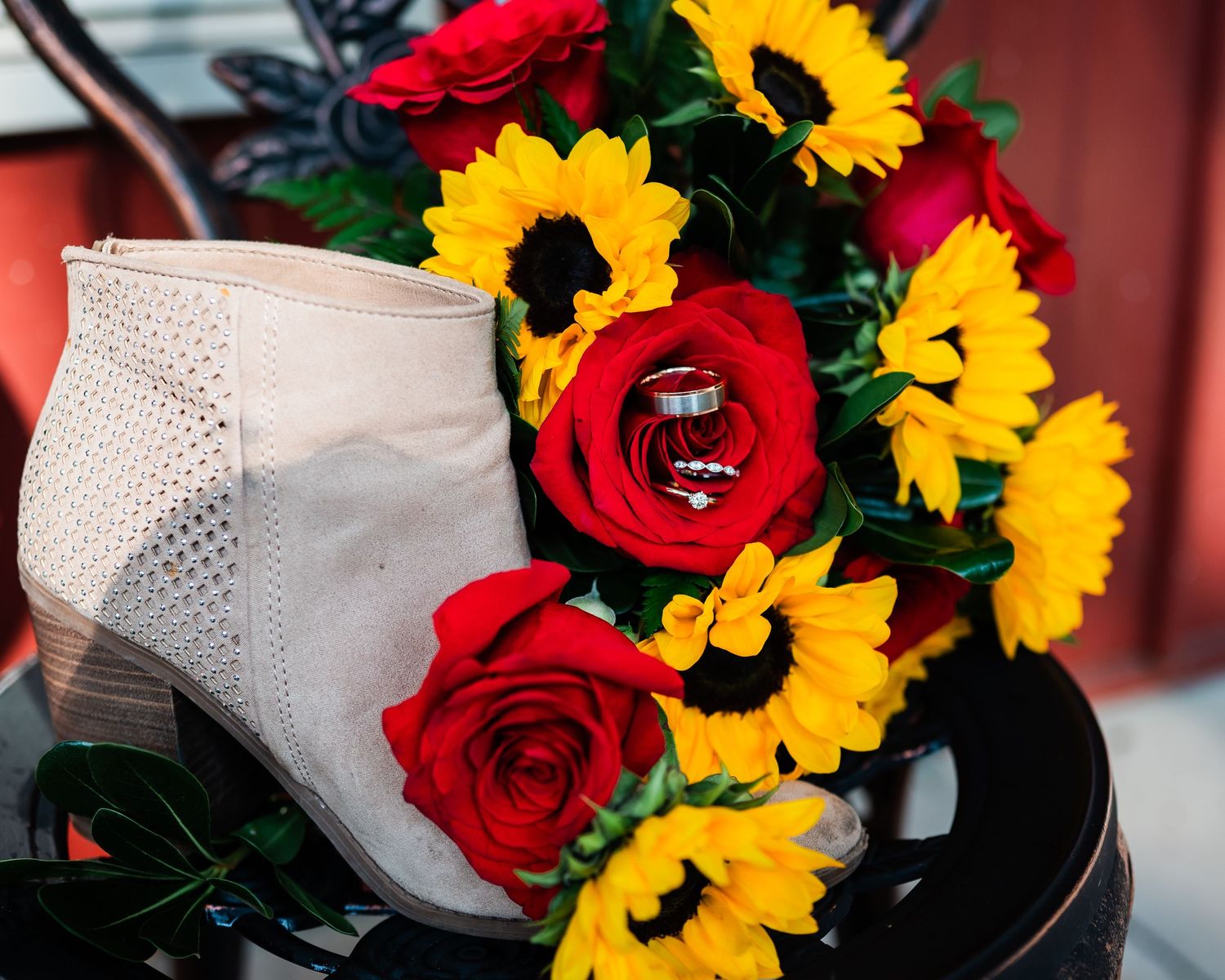 A bridal bouquet with bright red roses and yellow sunflowers next to a white lace boot.