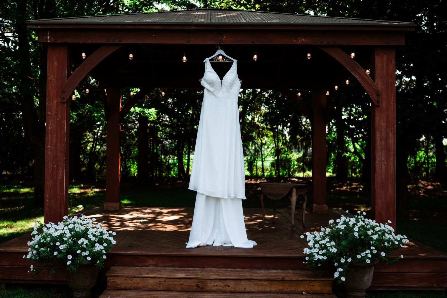 A white wedding dress hanging on a wooden gazebo decorated with white flowers.