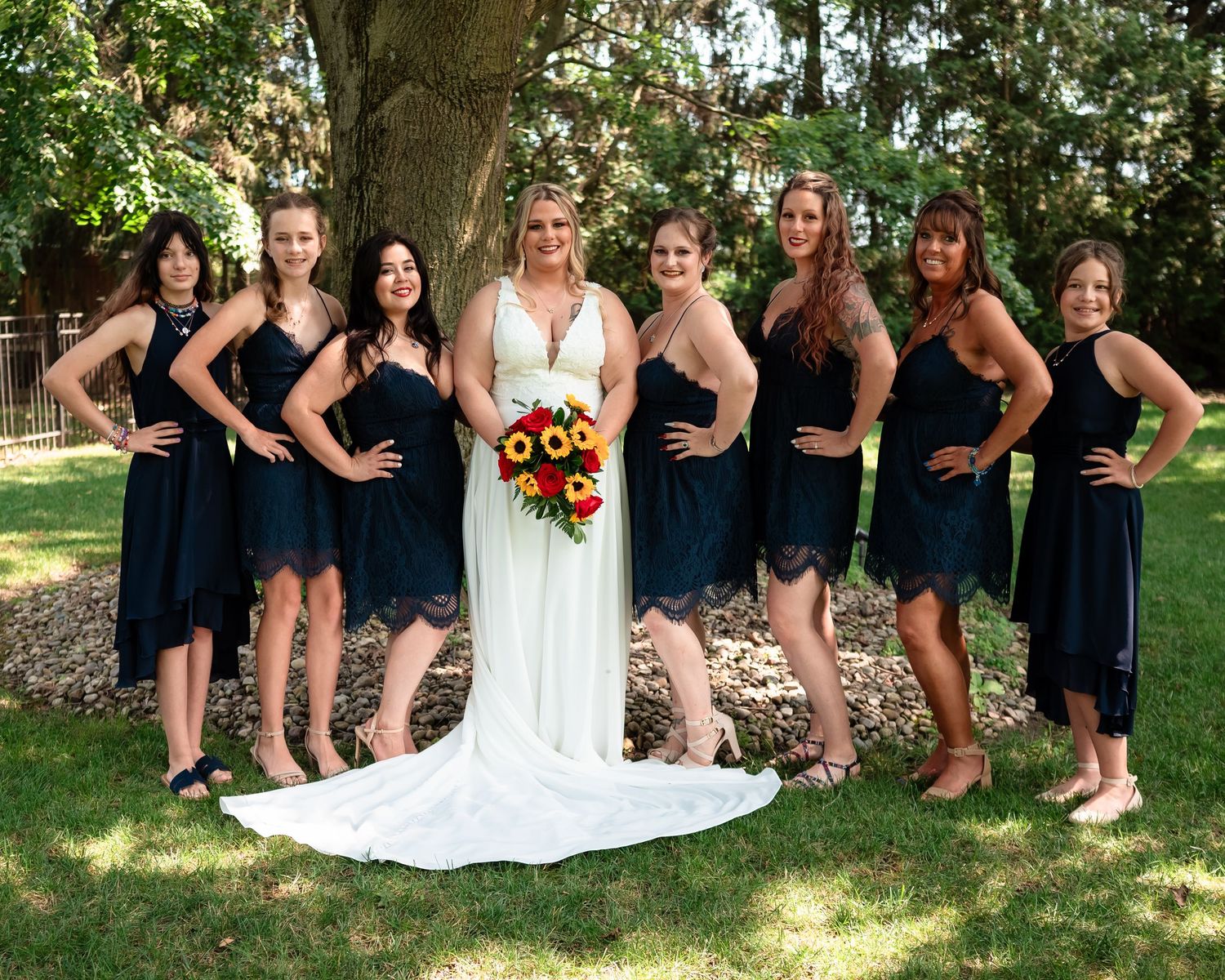 Wedding party photo with bridesmaids in navy dresses standing outdoors under a tree.