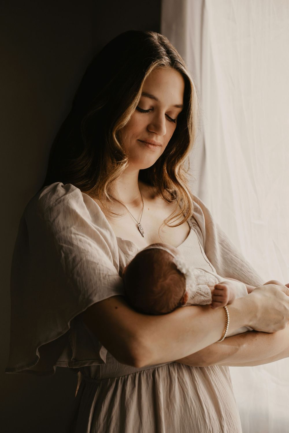 A mother in a cream dress holds and nurtures a newborn baby against natural window light in a motherhood photography session.