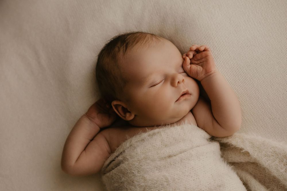Sleeping newborn baby wrapped in cream blanket with hand near face against neutral background.