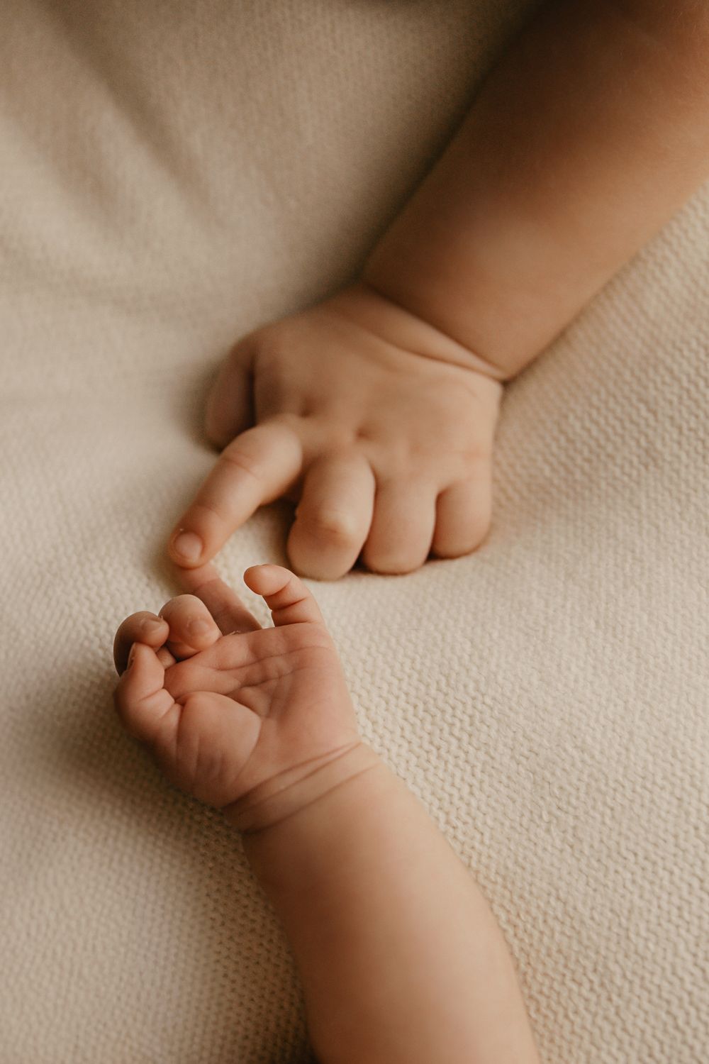 Close-up view of tiny baby hands resting on soft beige textured fabric in gentle natural lighting.