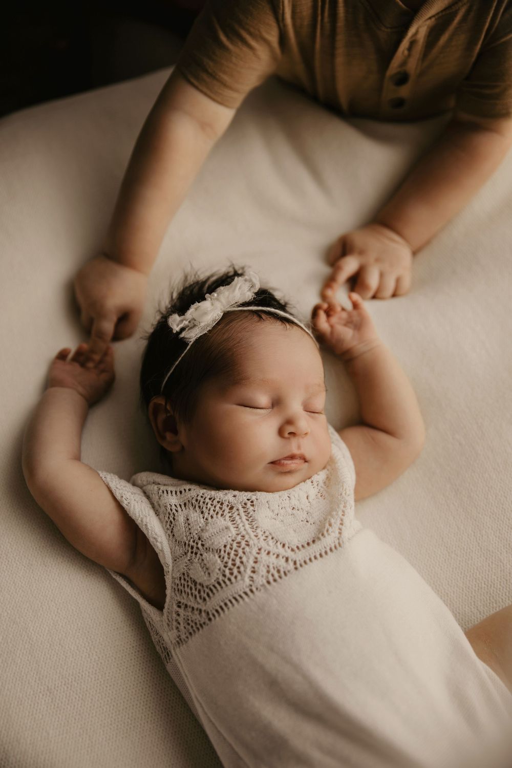 A sleeping newborn baby wearing a white lace dress lies peacefully on a cream-colored blanket.