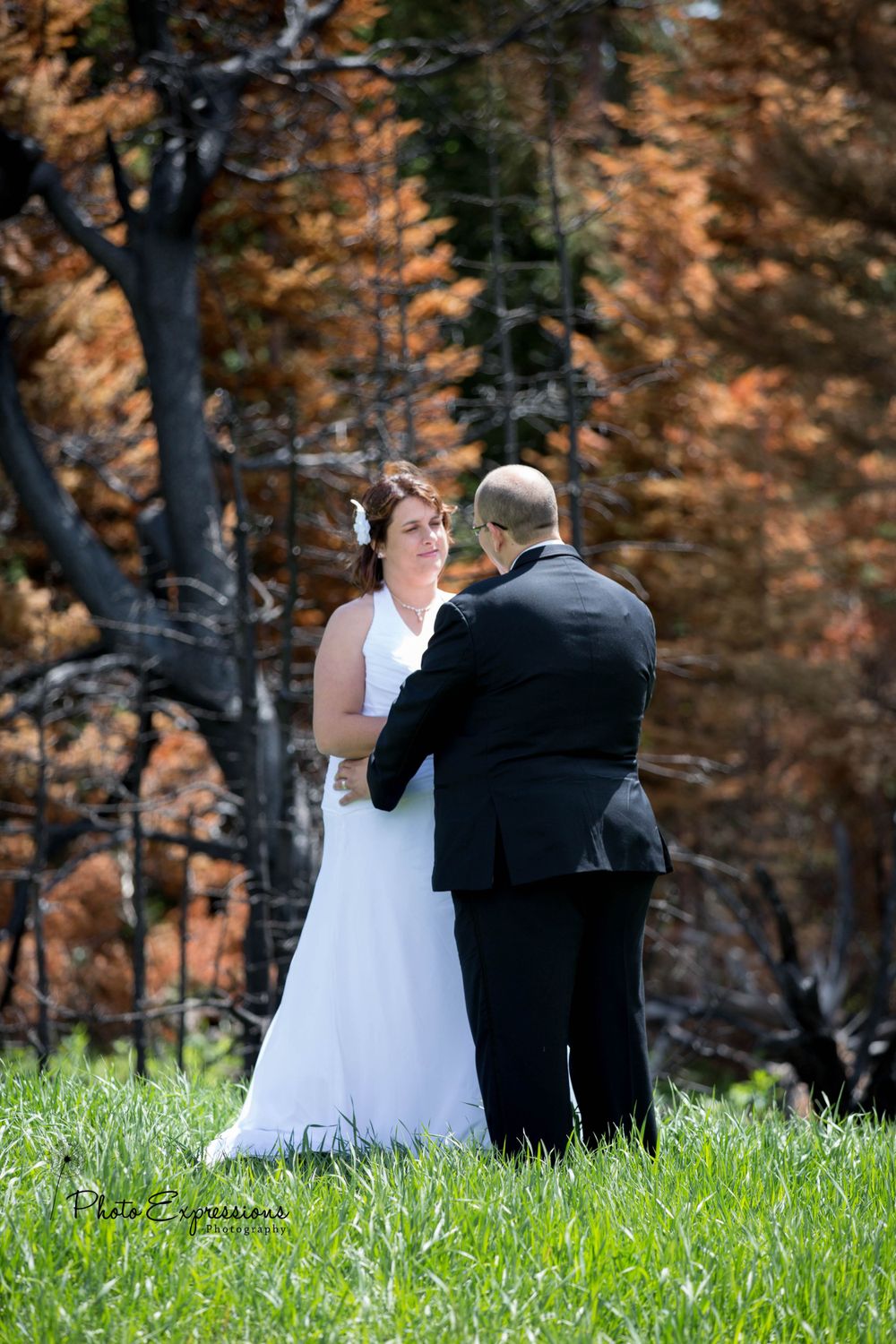Newlyweds sit together on grass with autumn foliage in the background.