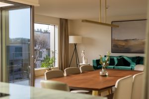 Modern dining room with wooden table, chairs and emerald green sofa near floor-to-ceiling windows.