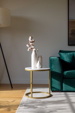 Minimalist side table with white vases holding cotton stems next to emerald green velvet armchair.