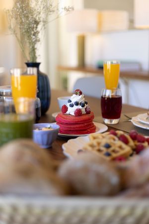 Red velvet pancakes topped with berries and cream beside orange juice and pastries.