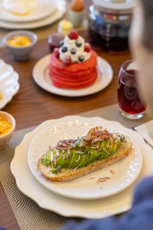 A gourmet sandwich with fresh greens on a white plate next to red pancakes topped with berries and cream.