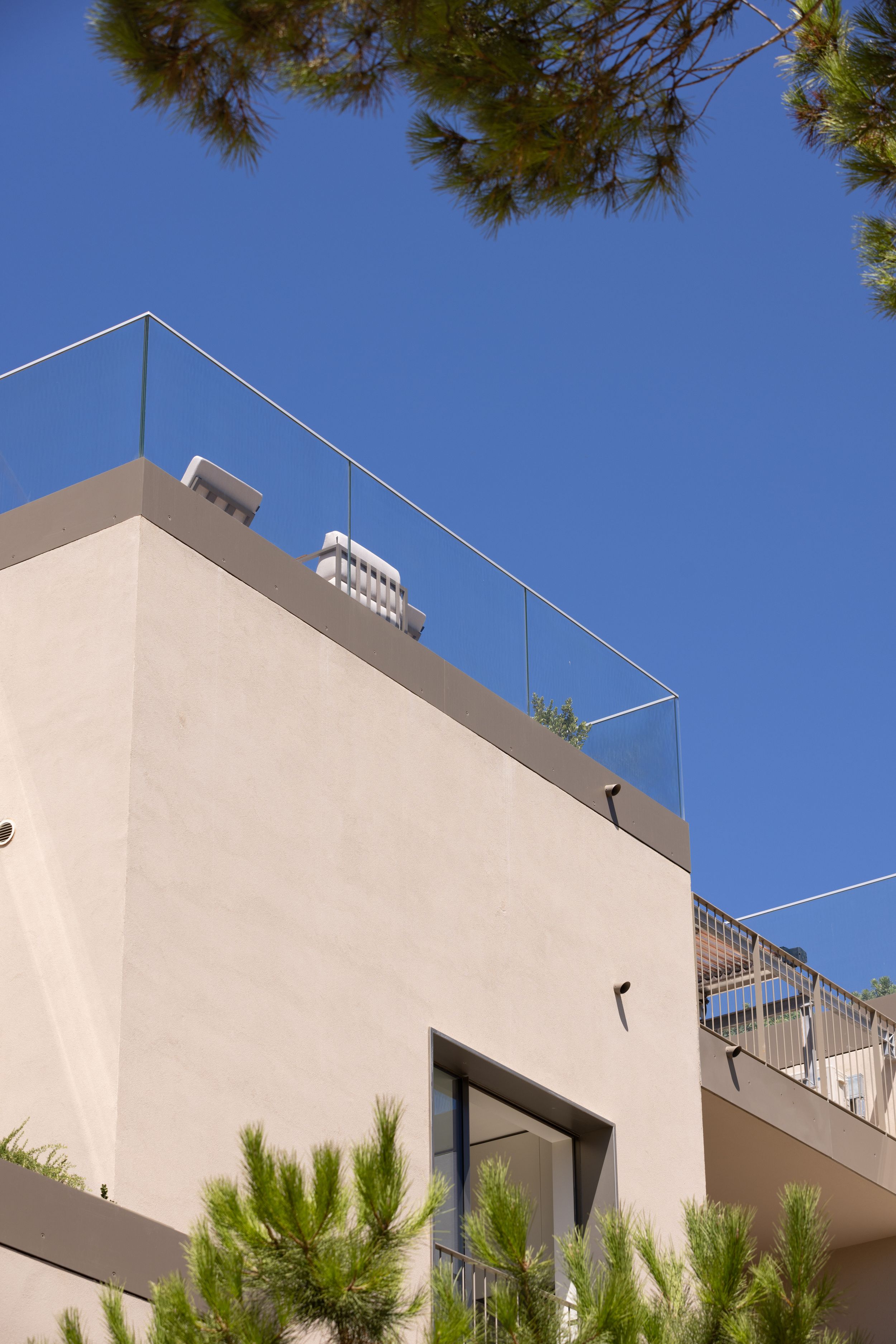 Modern architectural detail of a beige building with glass railing against blue sky and pine trees.