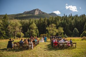Outdoor wedding ceremony in a meadow with mountain backdrop and evergreen forest under blue sky.