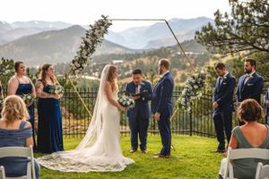 Outdoor wedding ceremony on grass overlooking mountain vista with floral arch and navy-clad wedding party.