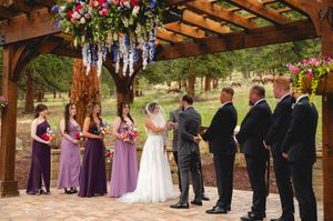 A wedding party stands outdoors in a line wearing purple and black formal attire beneath wooden beams decorated with flowers.