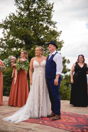 Wedding party stands on a red rug outdoors with bridesmaids in rust-colored dresses and elegant white wedding gown.