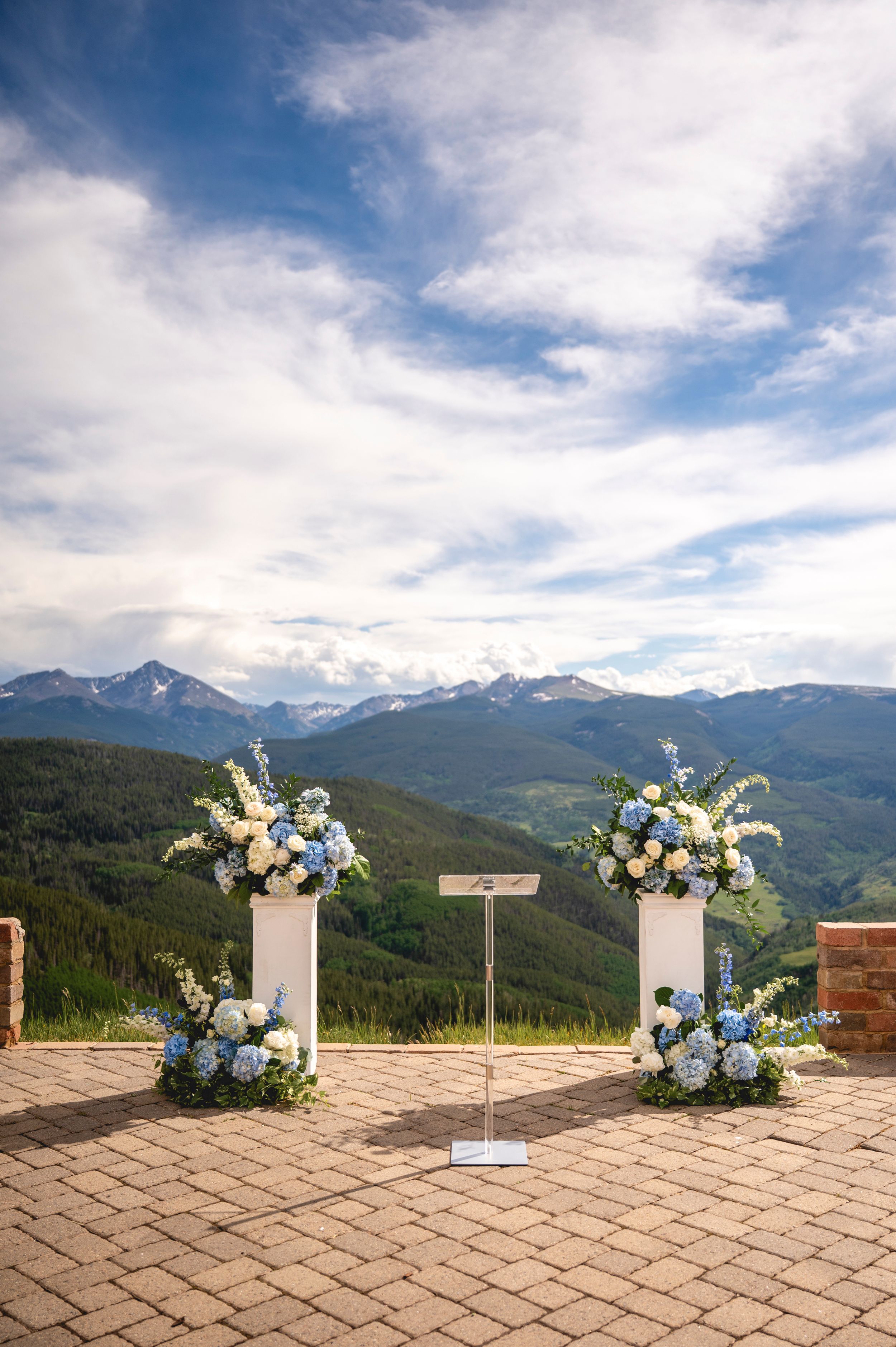 Outdoor wedding altar with blue and white floral arrangements on white pedestals, overlooking a stunning mountain range under a blue sky.