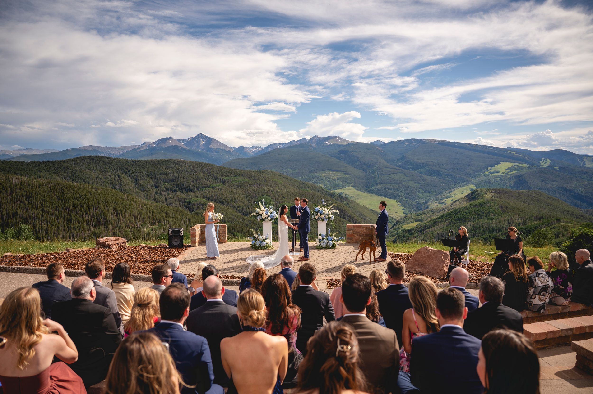 Outdoor mountain wedding ceremony with guests seated overlooking scenic Colorado Rocky Mountain peaks and lush green valleys.