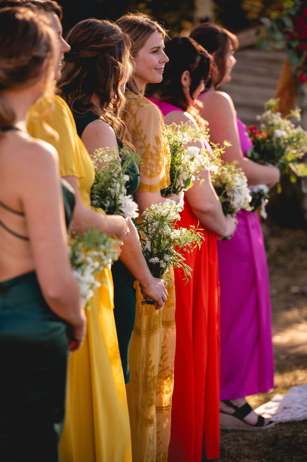 Bridesmaids in colorful rainbow dresses holding wildflower bouquets at outdoor ceremony.