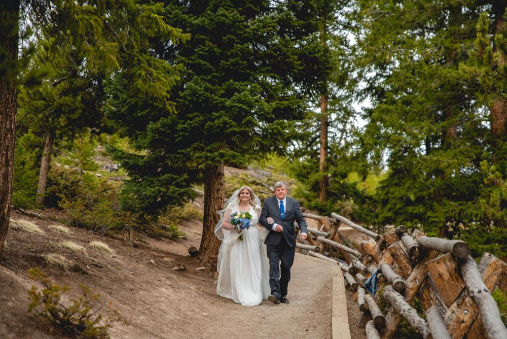 Couple walking down forest path surrounded by evergreen trees in mountain setting.