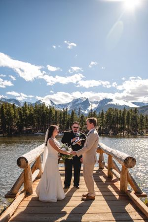 A wedding ceremony takes place on a wooden dock overlooking a mountain lake with snow-capped peaks in the background.