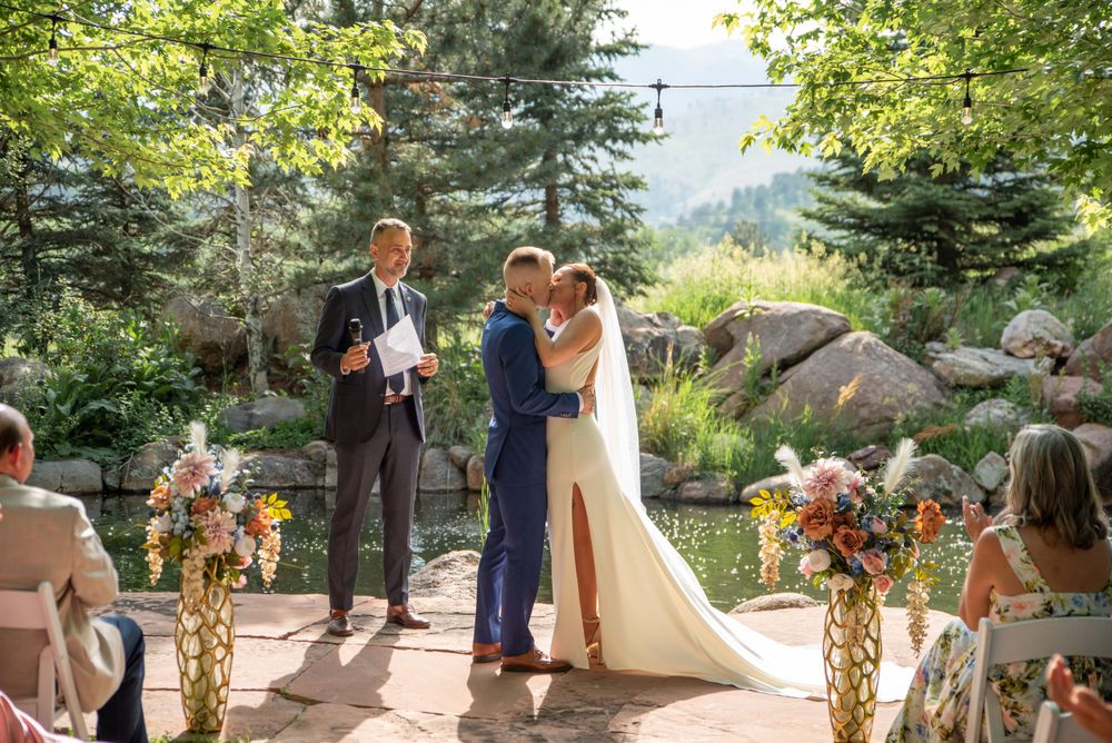 Outdoor summer wedding ceremony with floral decorations and mountain backdrop.