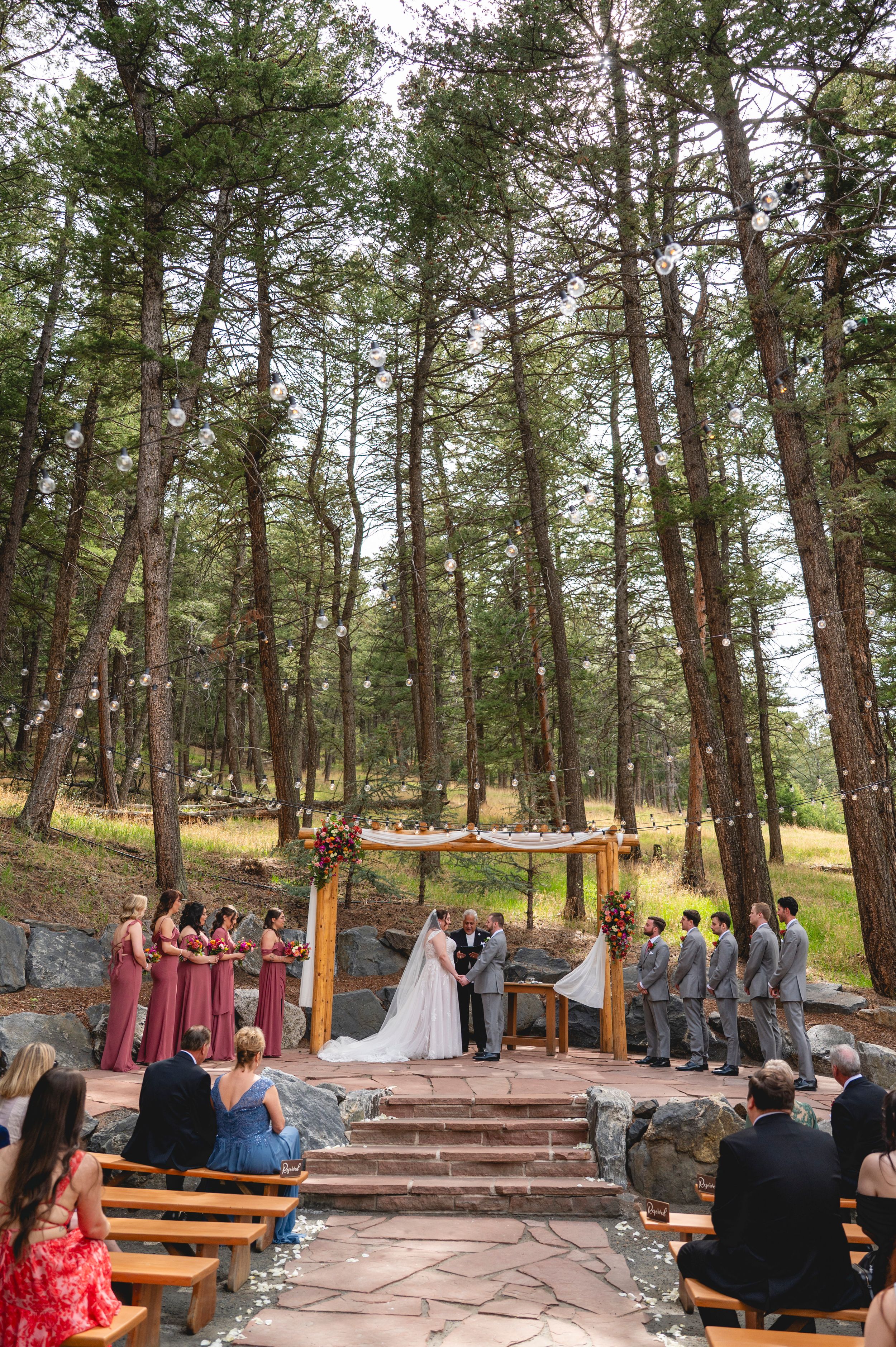 Outdoor forest wedding ceremony with bride and groom at floral arch, bridesmaids in mauve, groomsmen in gray, wooden pews.