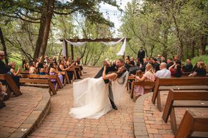 A wedding ceremony takes place in an outdoor wooded venue with wooden benches and white fabric decorations.
