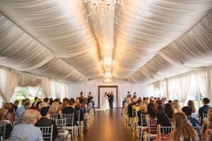 Elegant indoor wedding ceremony in draped white tent with crystal chandelier and rows of chiavari chairs.