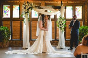 A wedding ceremony takes place in a rustic wooden venue decorated with white drapes and greenery under warm lighting.