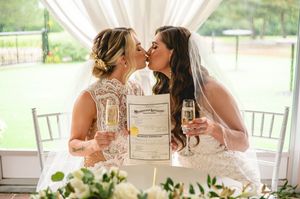 Two brides share a kiss during their wedding celebration while holding champagne glasses and a marriage certificate.