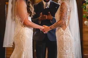 Two people in lace wedding dresses hold hands during a ceremony as an officiant stands in the background.
