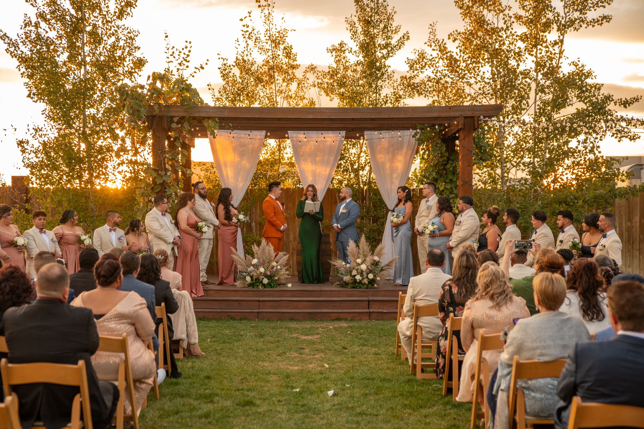 Golden sunset wedding ceremony under wooden pergola with rows of guests seated on wooden chairs.