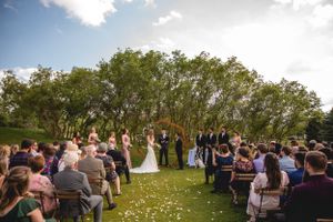 Garden wedding ceremony with guests seated on white chairs under trees and scattered flower petals.