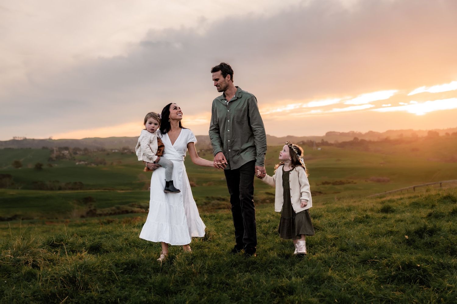 Family standing together in a green field during golden sunset with rolling hills in background.