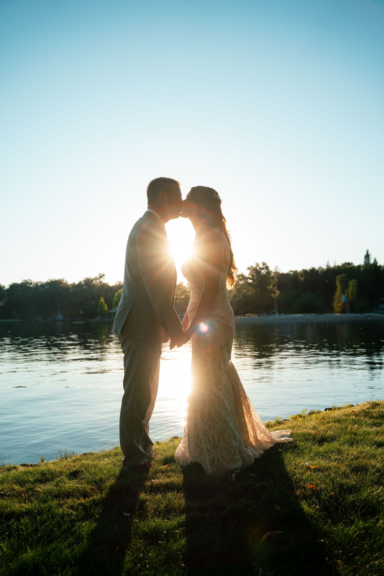 Silhouetted couple embraces by a tranquil lakeside at sunset, creating a romantic backlit scene against the water.