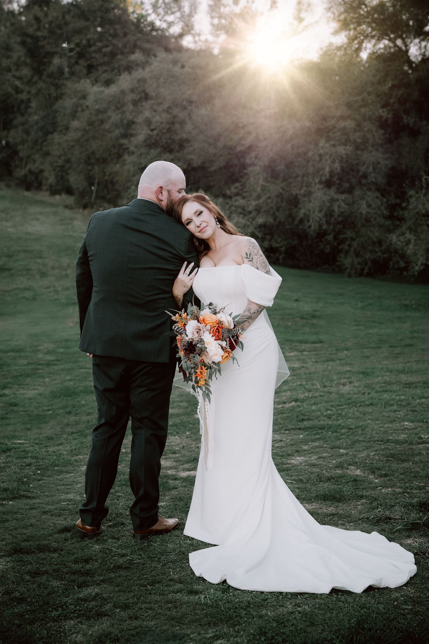 A romantic wedding couple shares a tender moment at sunset in a grassy field with warm golden light.