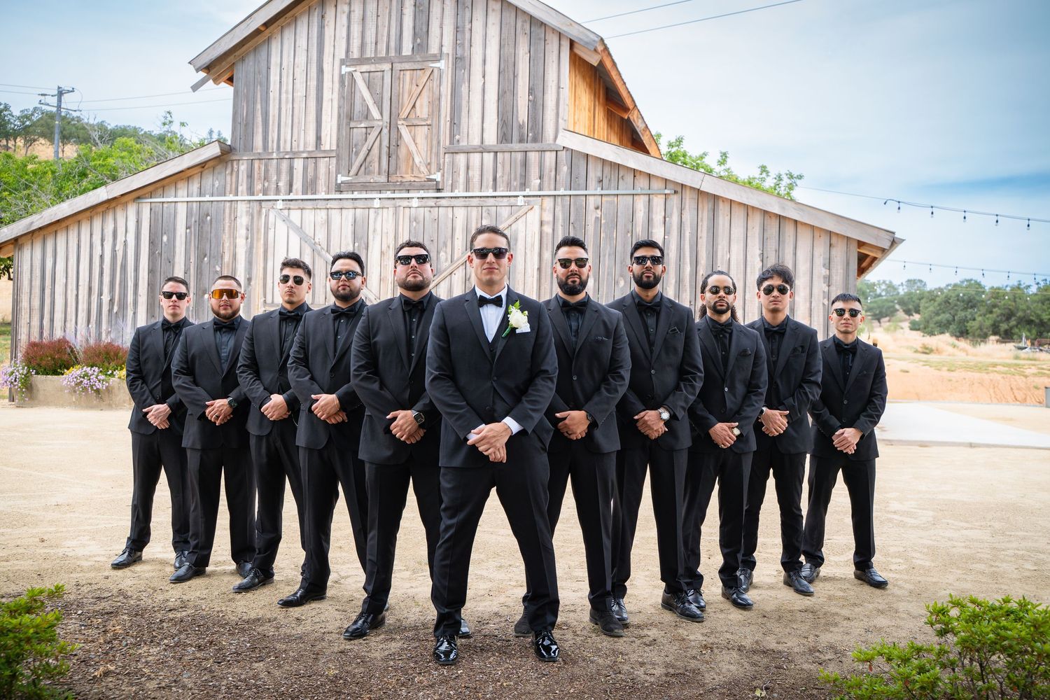 Wedding party wearing black suits and sunglasses pose in V-formation outside rustic wooden barn.