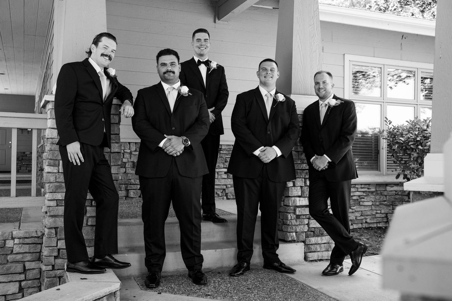 Five groomsmen in black tuxedos pose together on stone steps outside a building in a classic black and white wedding photo.