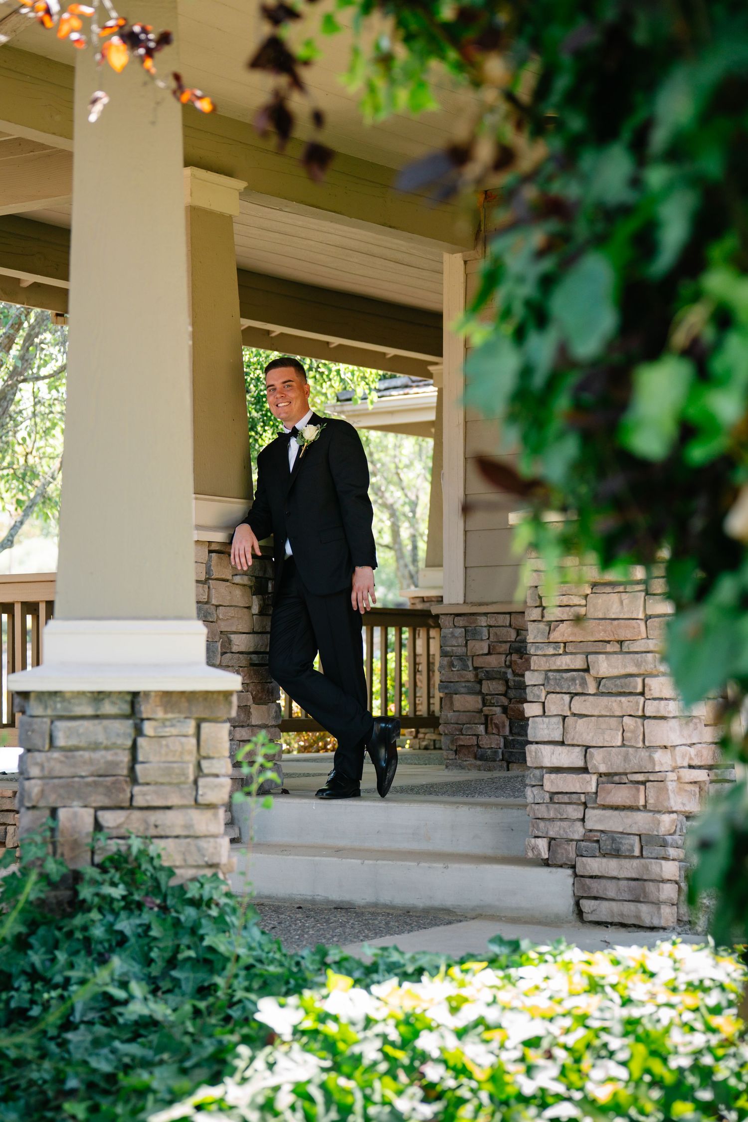 Groom in black tuxedo and bow tie standing on stone porch steps surrounded by lush greenery.