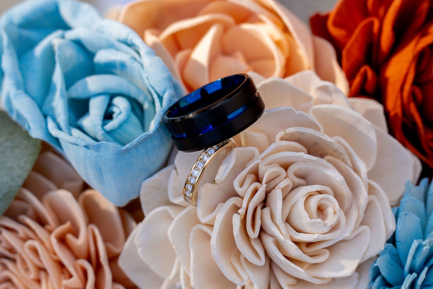 Blue and black wedding band with diamond engagement ring resting on colorful paper flowers in blue, peach, and orange hues.