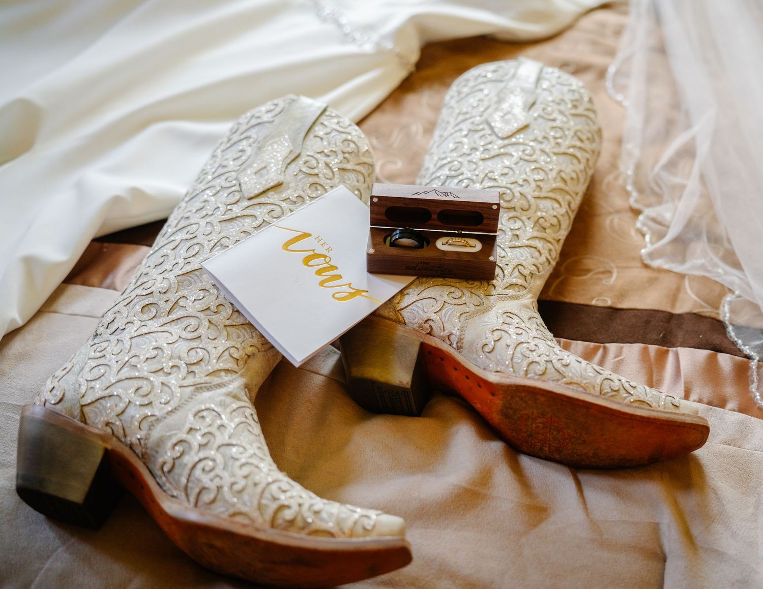 White embroidered cowboy boots with gold vow cards and a wooden ring box on a wedding day flat lay.