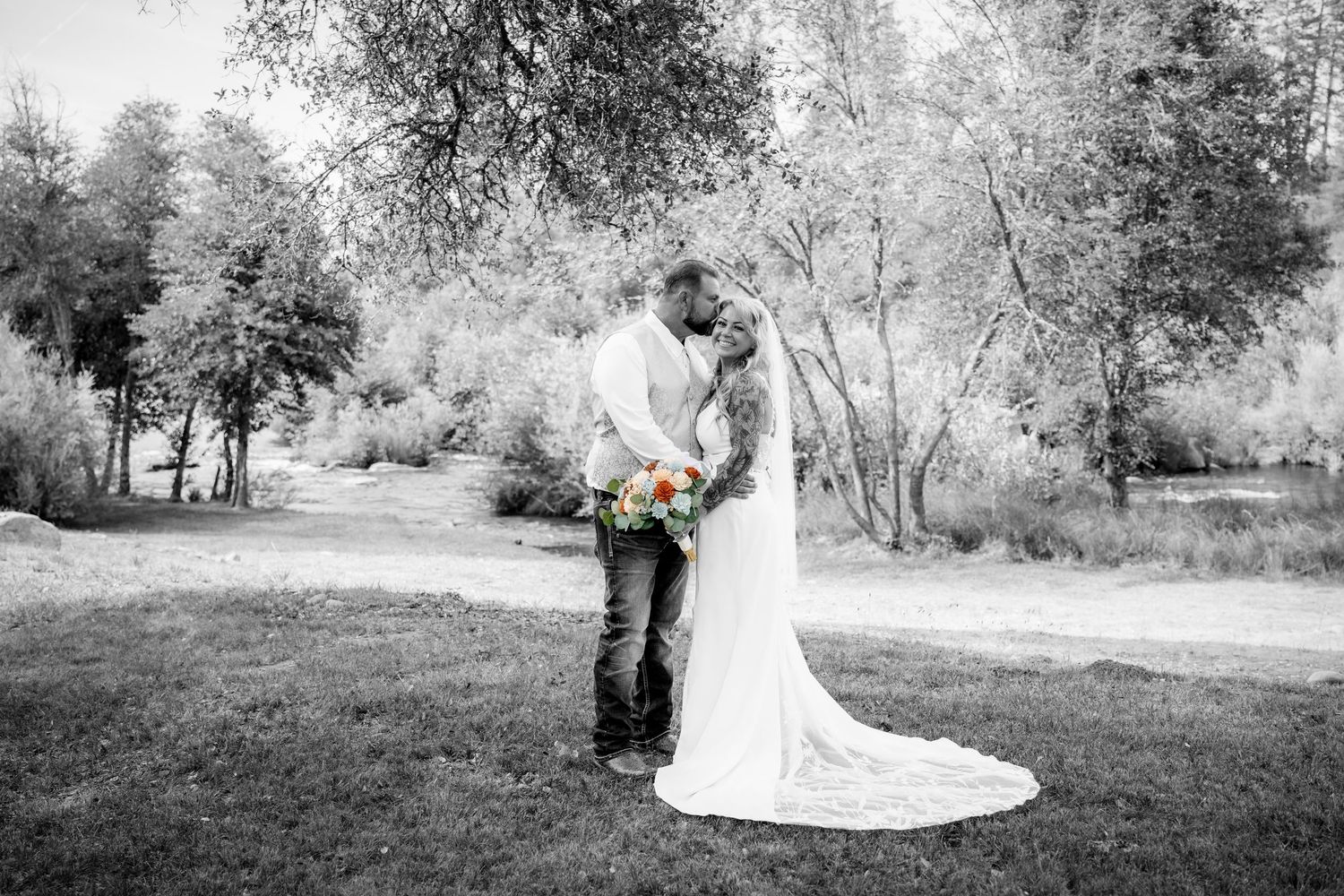 Black and white photo of a bride and groom kissing outdoors, bride holding colorful bouquet with trees in background.