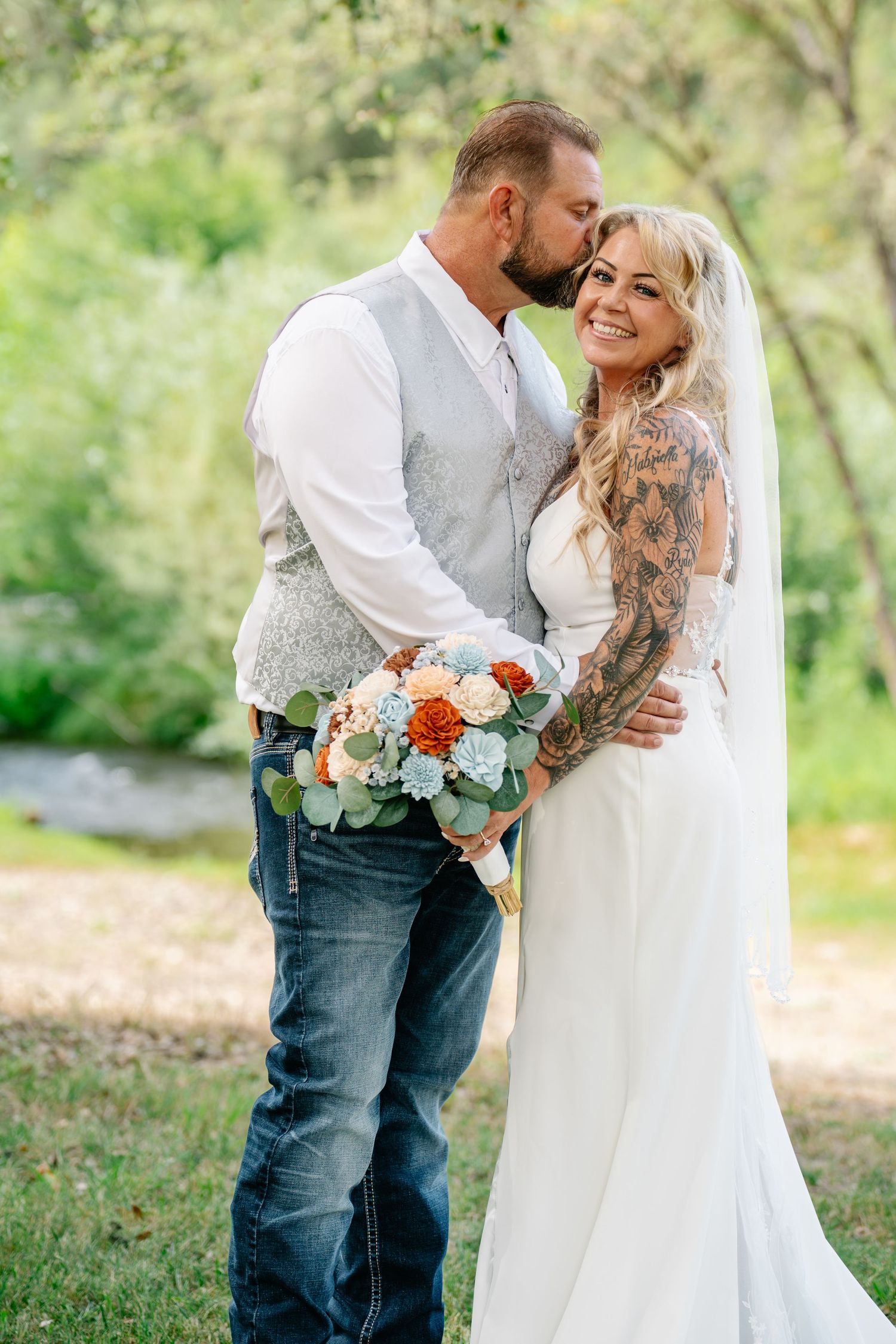 Bride with tattoos and groom in jeans share a tender moment outdoors, holding an orange and white bouquet.