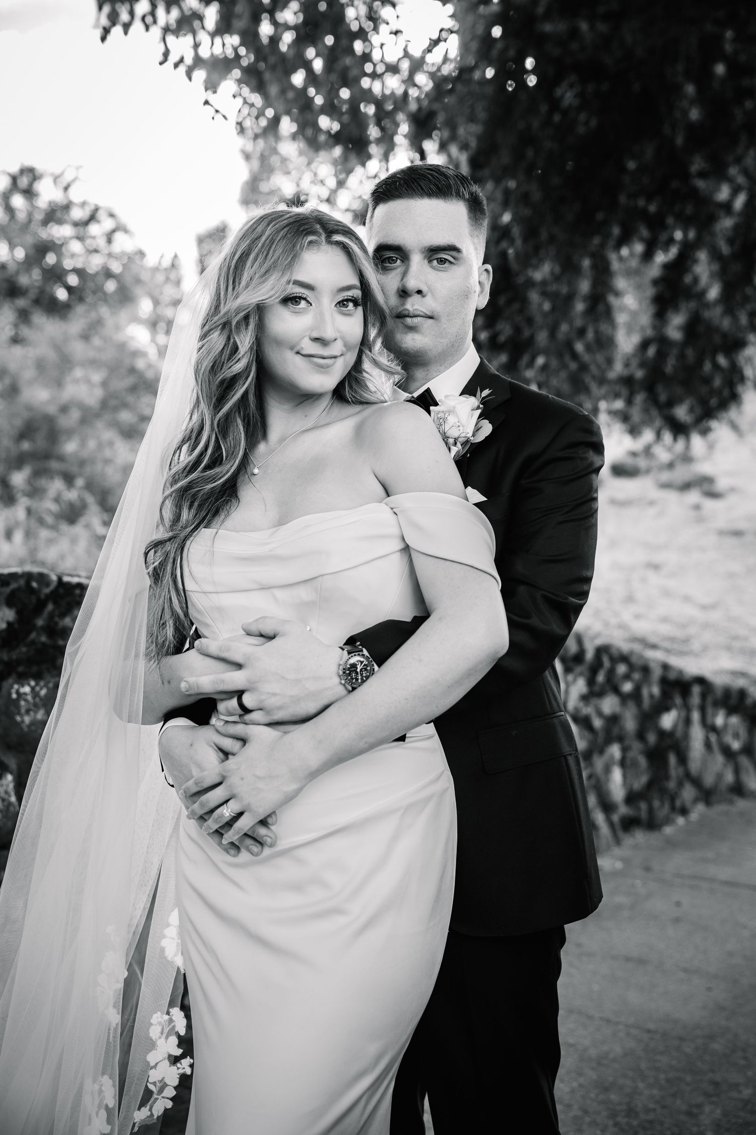 Black and white wedding portrait of bride in off-shoulder gown with veil and groom in dark suit embracing outdoors.