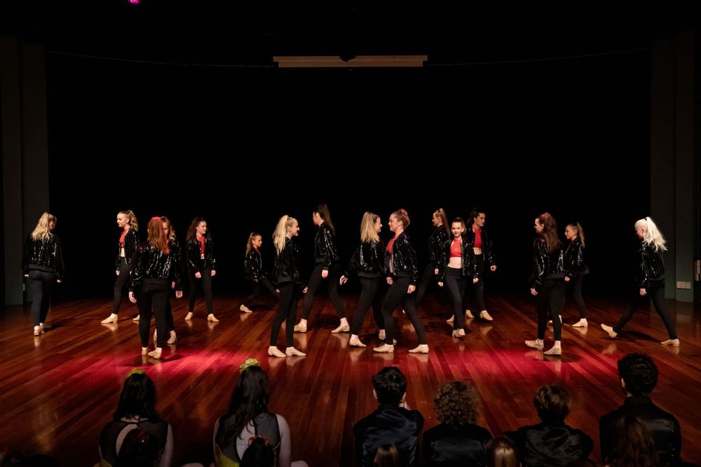 Dance group performs synchronized choreography on illuminated wooden stage with audience silhouettes.