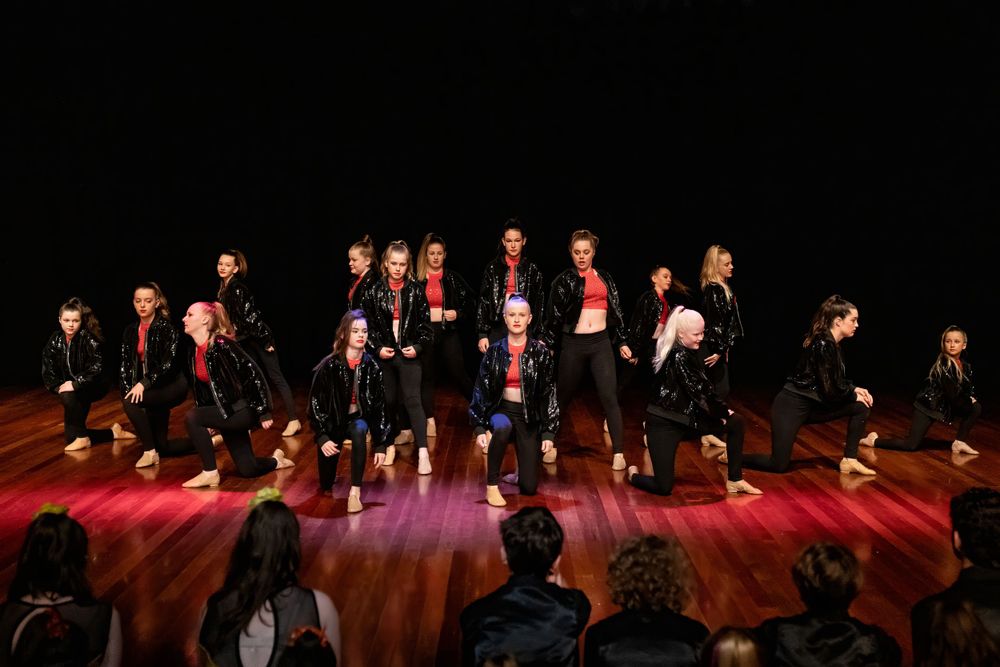 Dance ensemble in matching outfits performs on polished wooden stage with dramatic lighting.