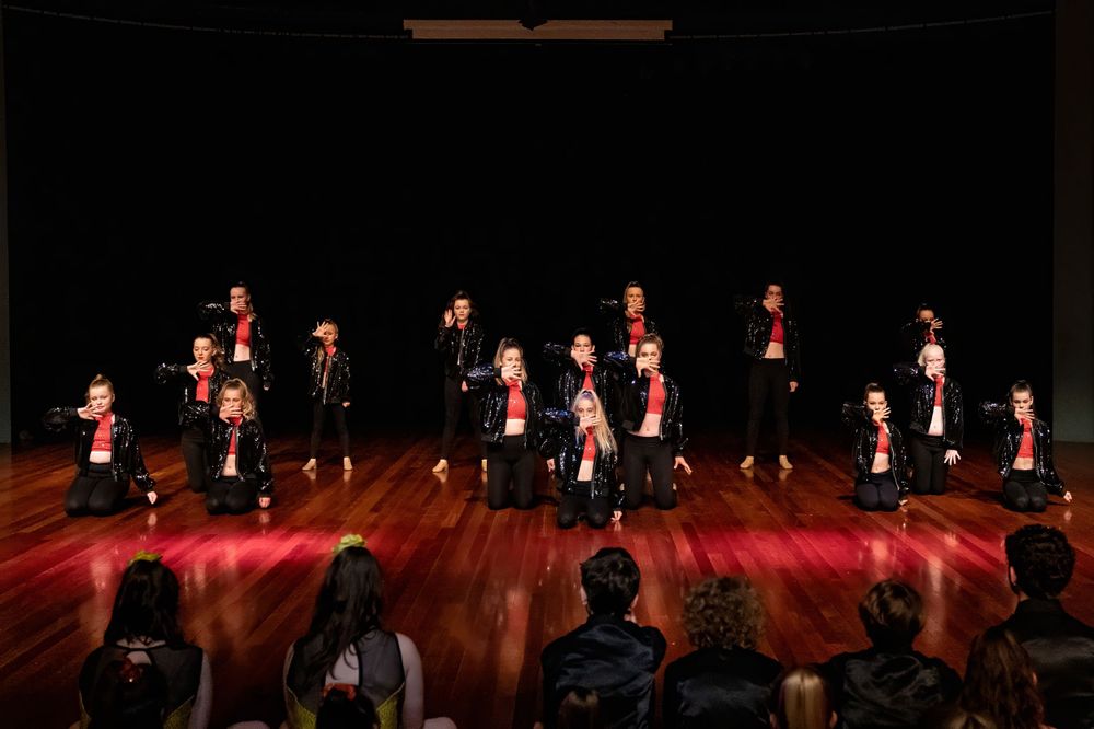 Performance group in black and red costumes arranged in formation on dark wooden stage.