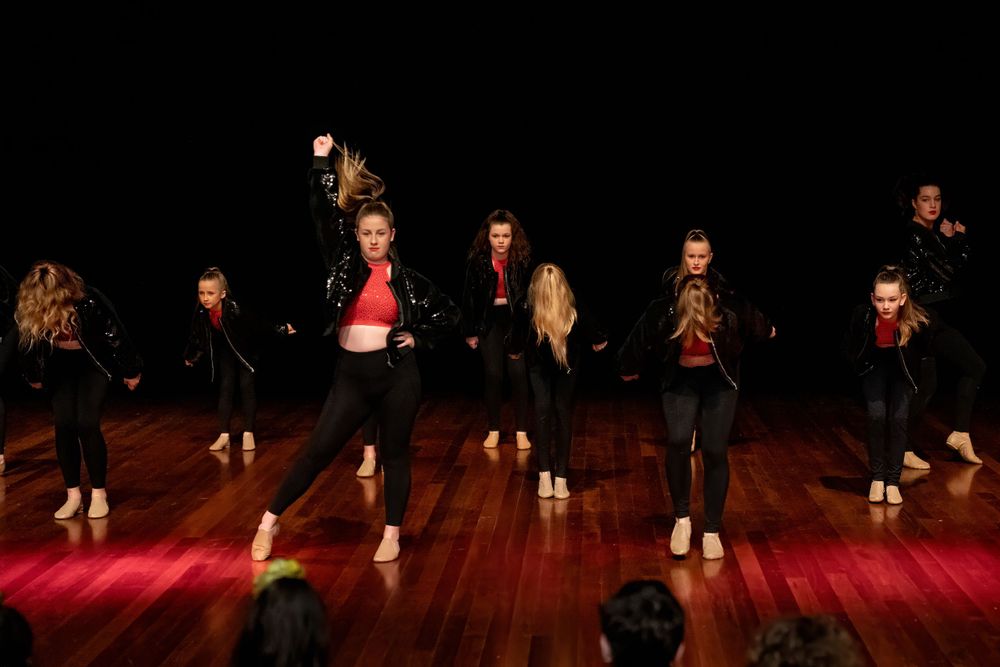 Dancers perform in black outfits on a wooden stage with dramatic red lighting.