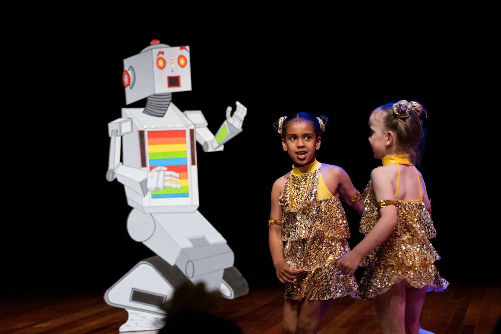 A robot character interacts with dancers in gold costumes on a dark stage.