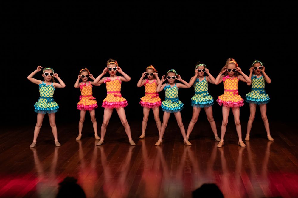 Eight dancers in colorful ruffled costumes perform synchronized poses on stage.