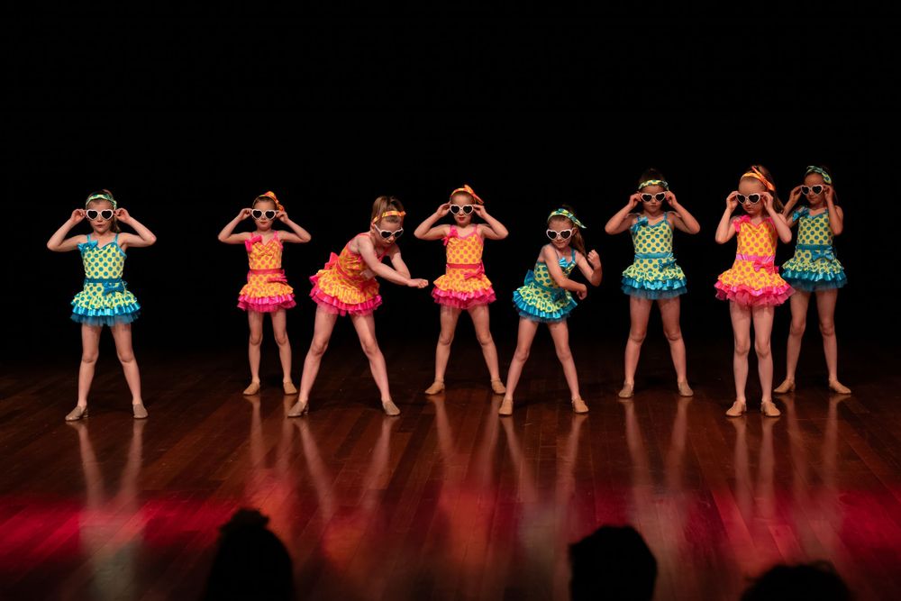 Line of dancers in vibrant tutus perform synchronized movements on wooden stage.