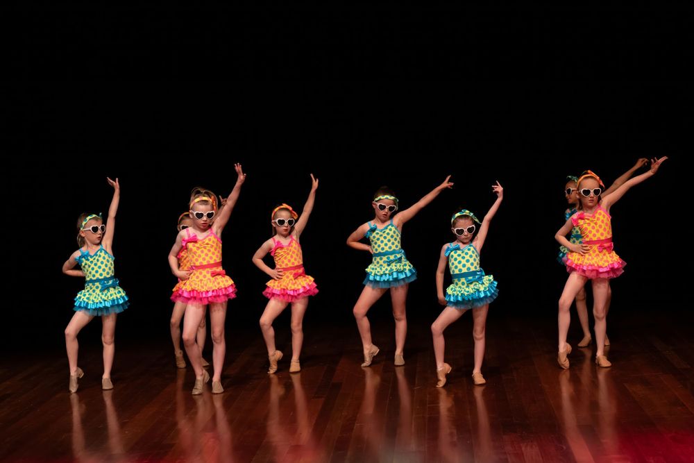Six dancers in colorful tutus perform expressive arm movements in stage formation.
